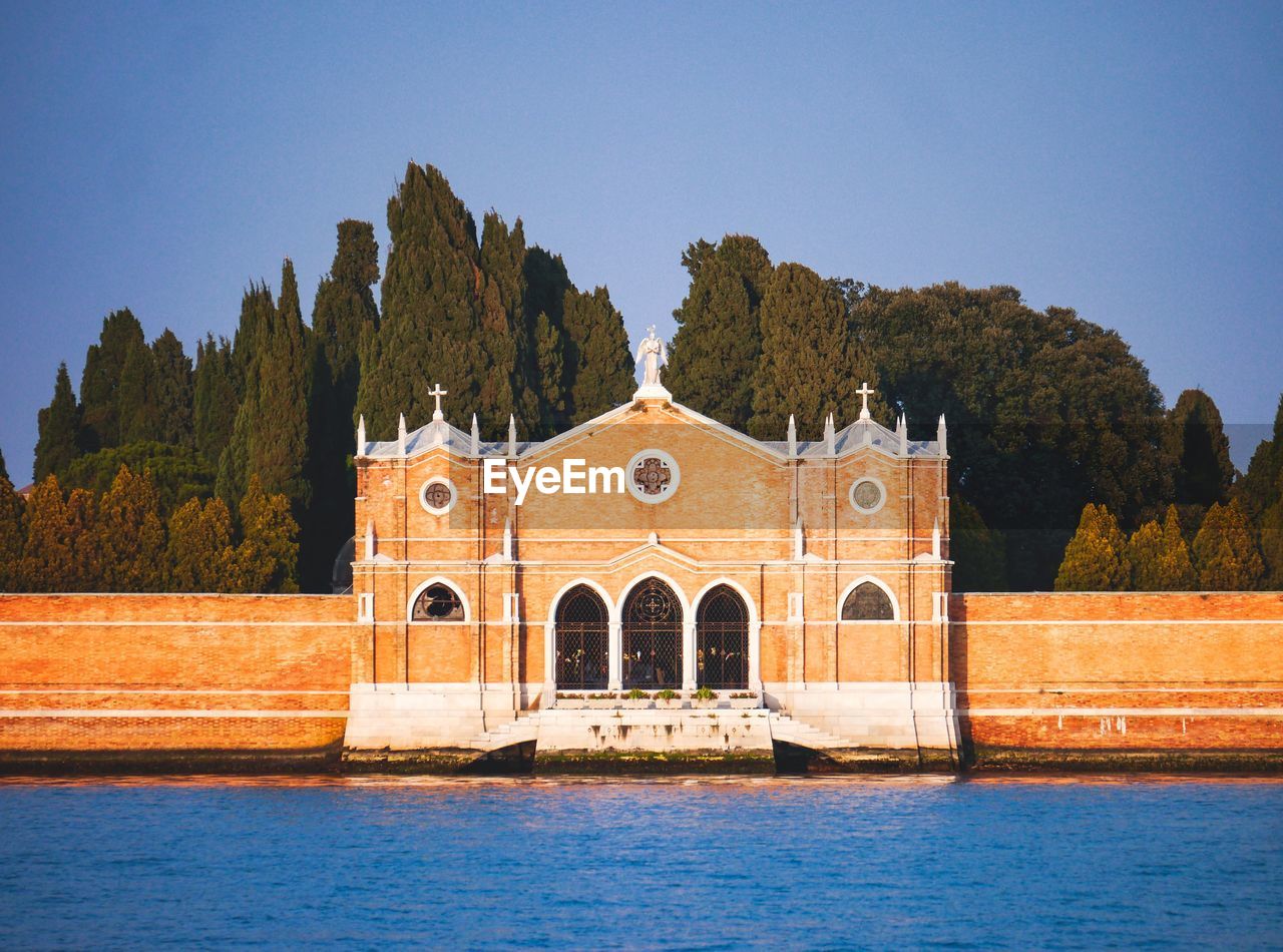 Building by river against clear blue sky - venice cemetery