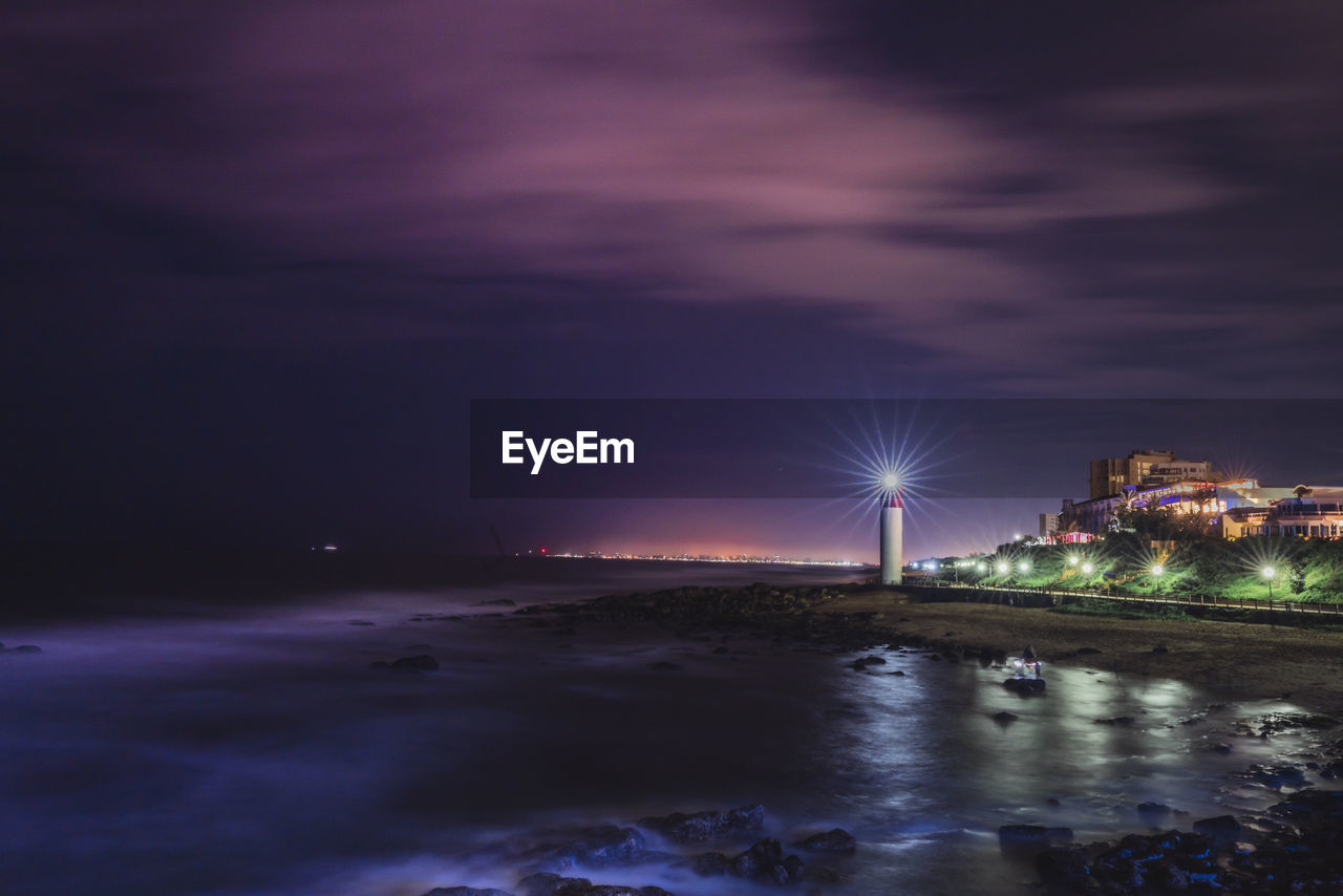 Illuminated city by sea against sky at night