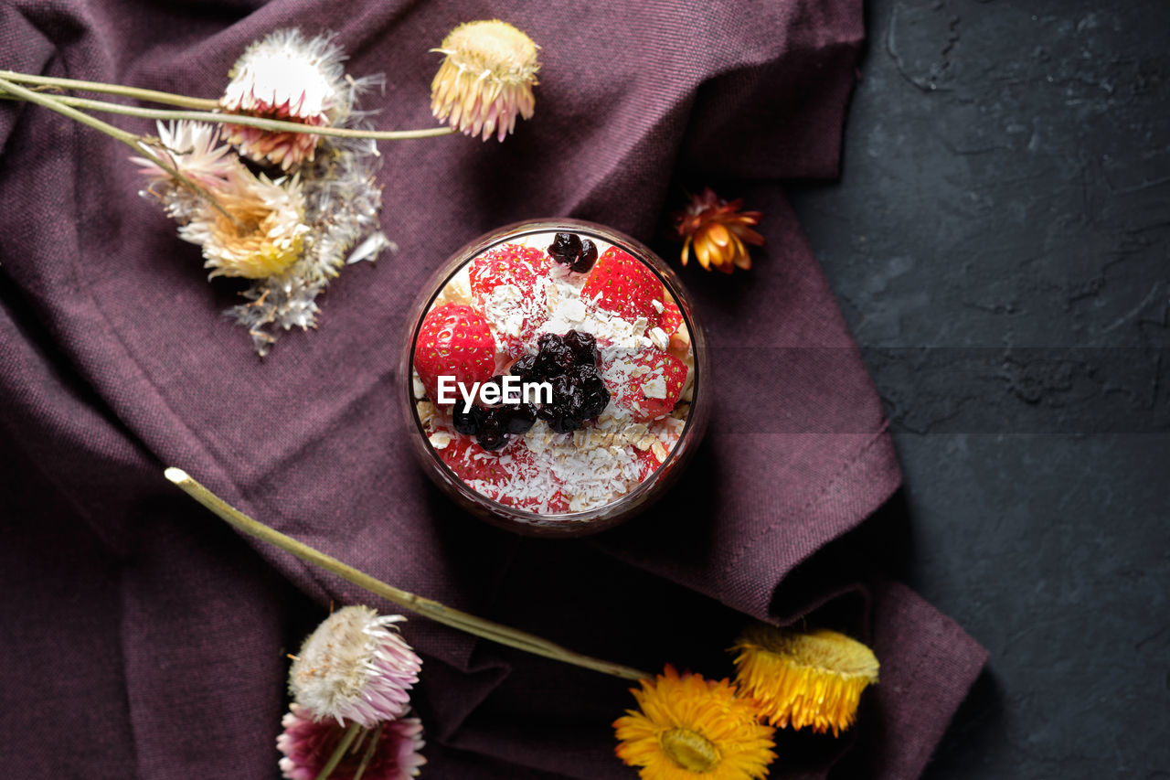 Top view appetizing smoothie bowl with granola strawberries and blueberries placed on table near various wildflowers