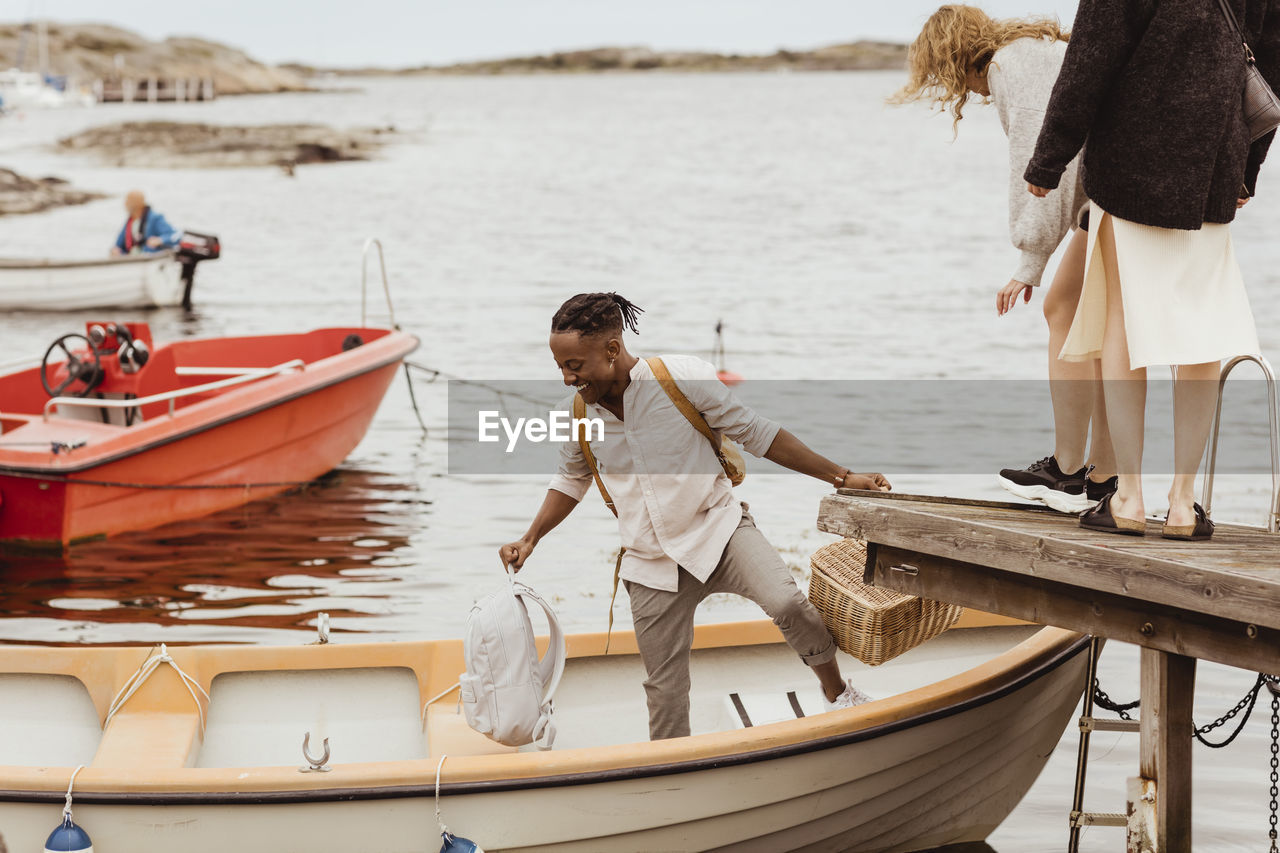 Smiling man helping female friends boarding boat by harbor