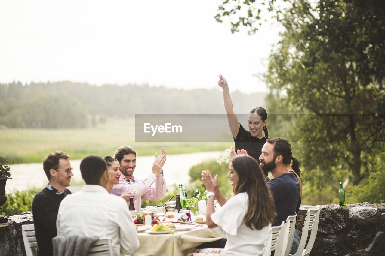 Mid adult woman standing with arm raised while friends applauding at table during dinner party in backyard