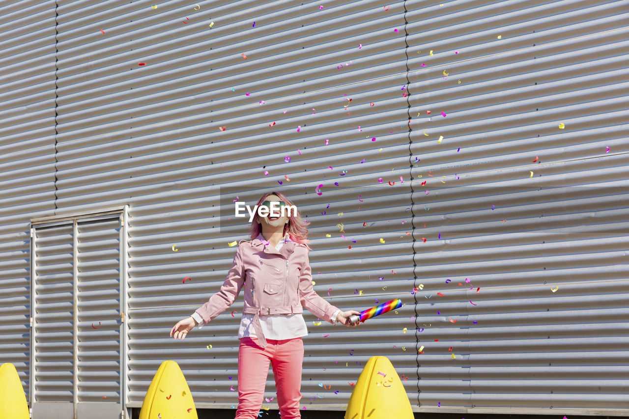 Cheerful woman with party popper having fun while falling confetti during sunny day