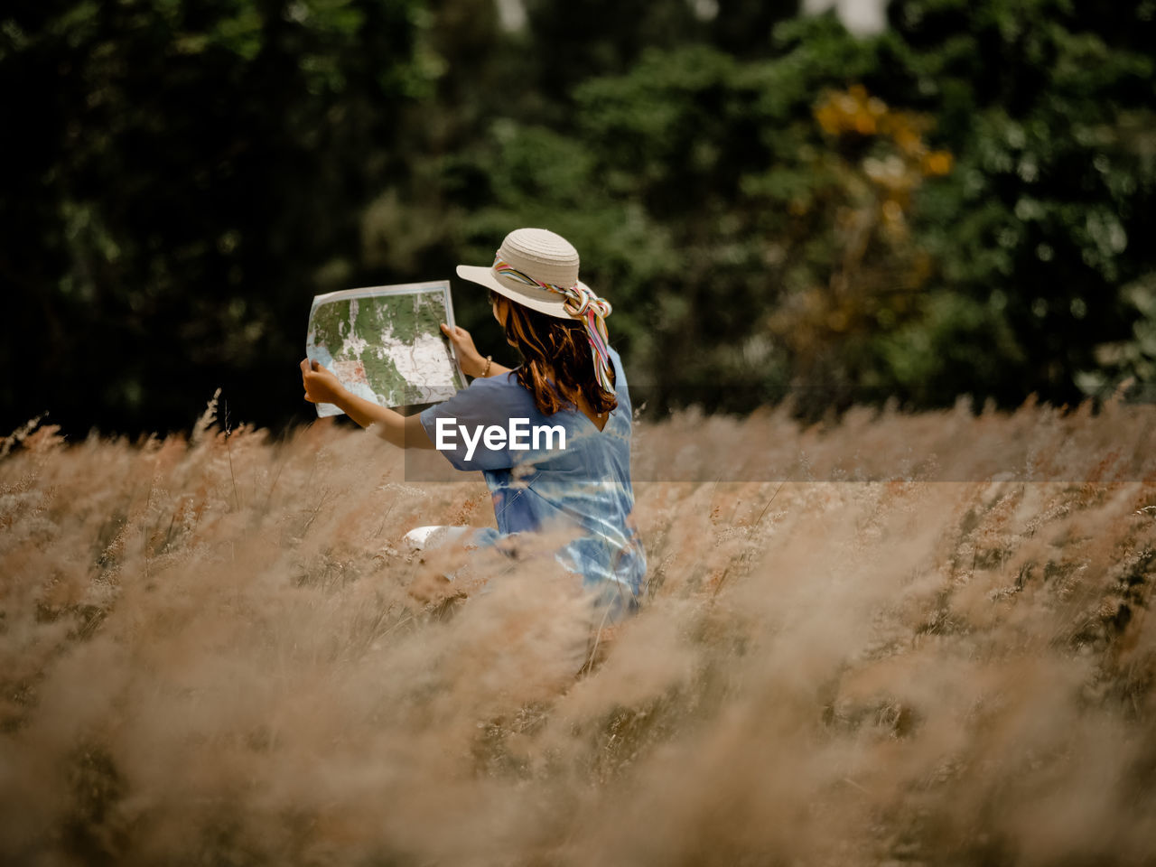 Woman wearing hat while reading map on field against trees