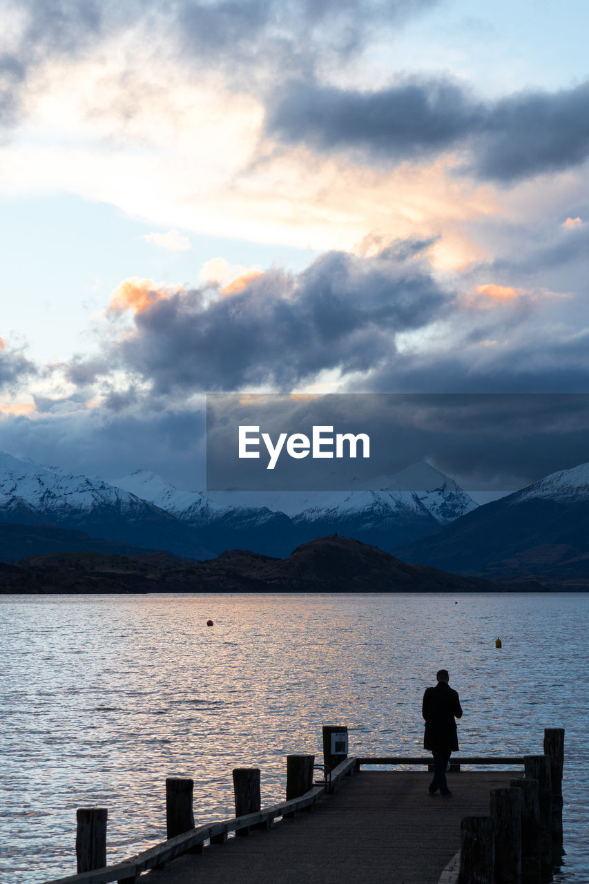 Man standing on pier by lake against sky during sunset