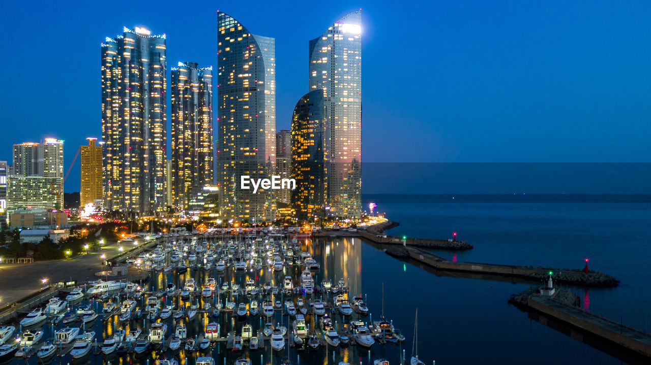 ILLUMINATED MODERN BUILDINGS BY SEA AGAINST SKY AT NIGHT IN CITY