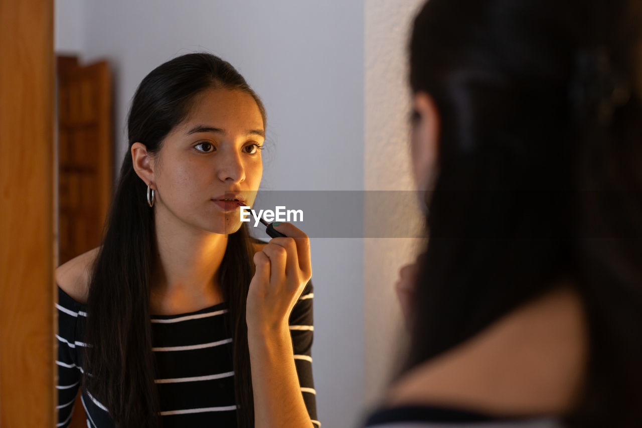 Young ethnic lady looking in mirror while applying lip gloss