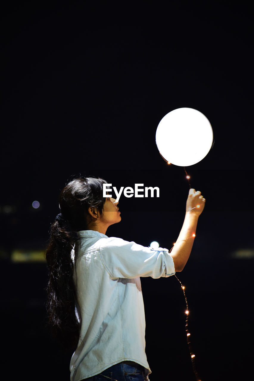 Woman holding illuminated balloon against sky