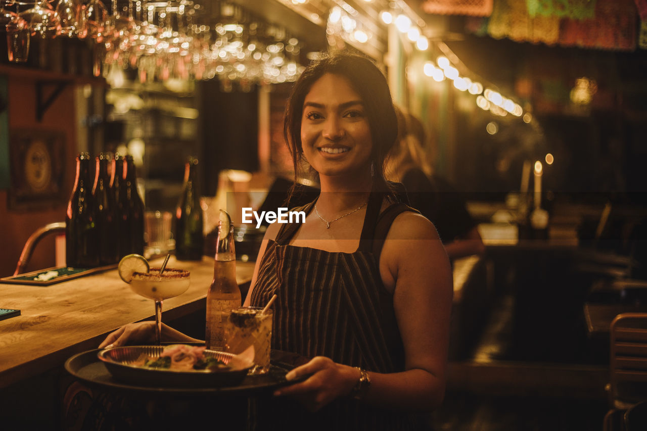 Portrait of smiling waitress holding food and drinks on serving tray while working at bar