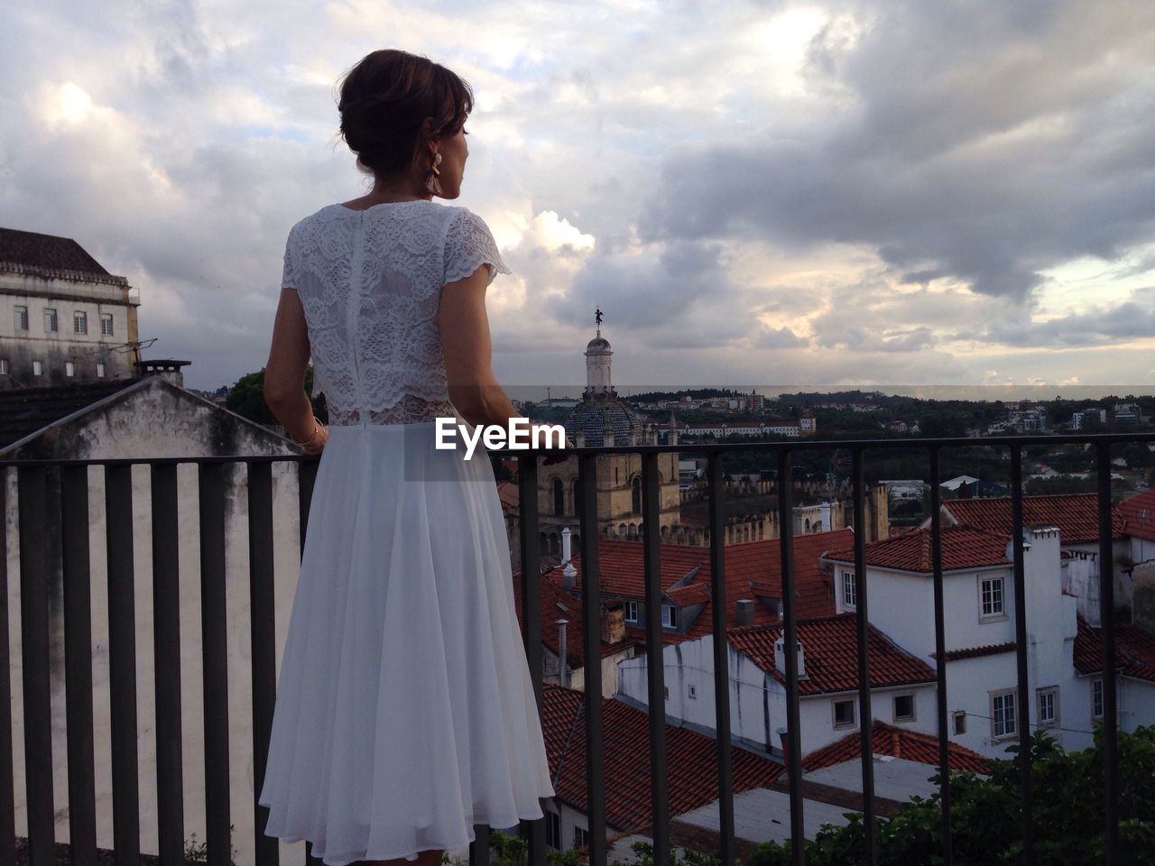 Rear view of young woman standing in balcony overlooking townscape against cloudy sky
