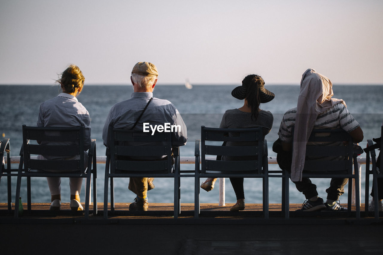 REAR VIEW OF PEOPLE SITTING ON CHAIR AT SEA SHORE