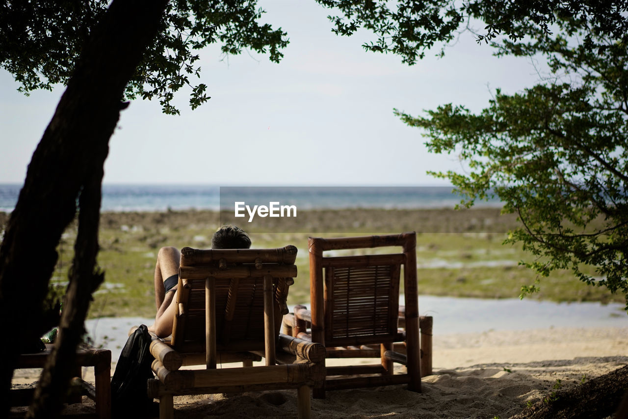 Rear view of man sitting on lounge chair at beach against sky