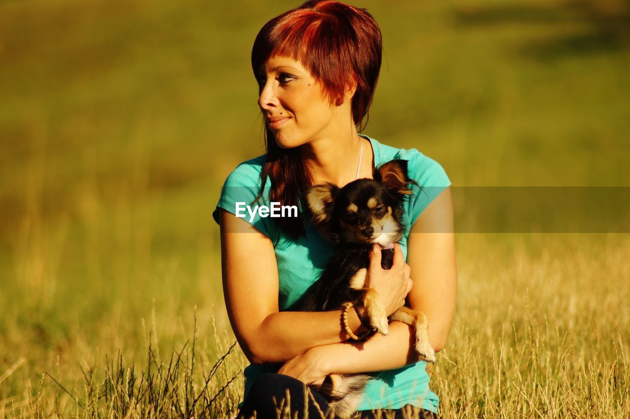 Smiling redhead woman carrying dog while sitting on grassy field