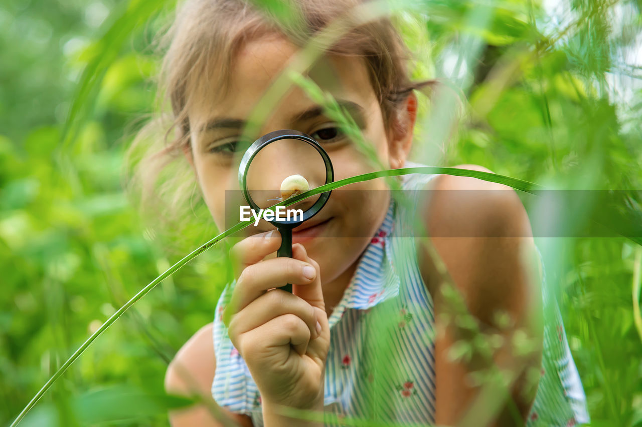 Front view of smiling girl looking at snail on grass through magnifying glass