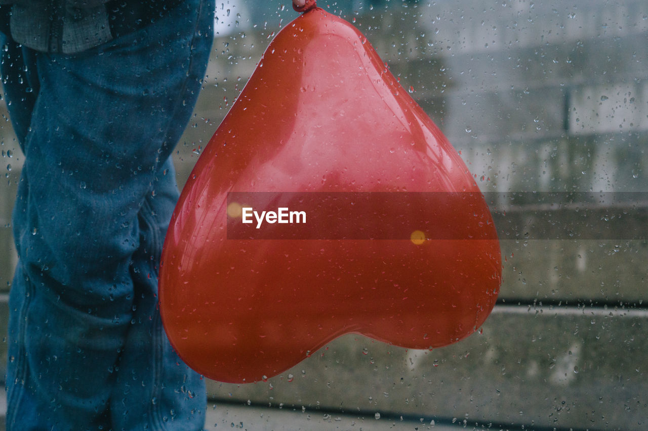 Midsection of person with heart shape balloon seen through wet glass during rainy season