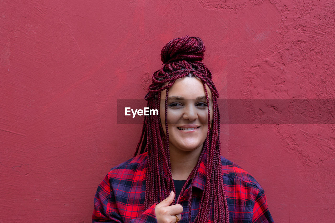 Portrait of young woman with dreadlocks standing against bright red wall