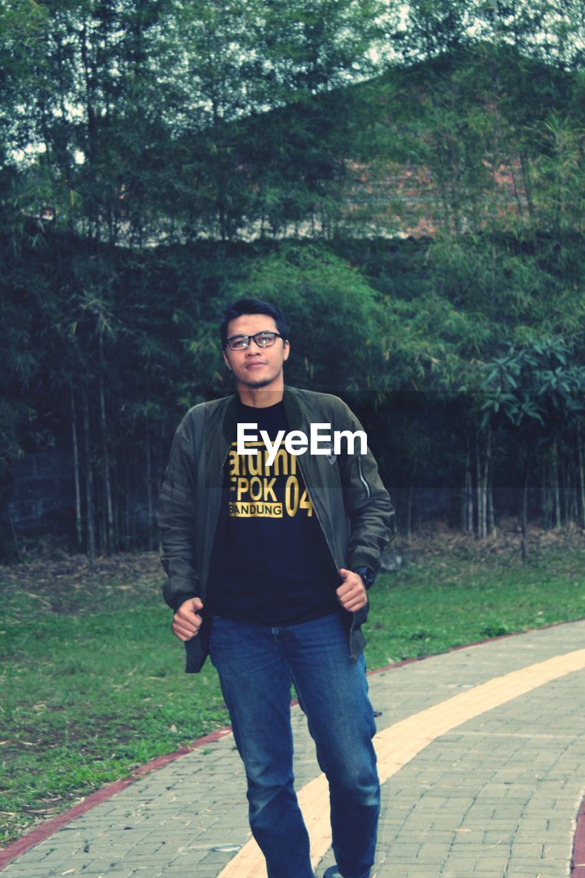 Portrait of young man standing against trees on footpath