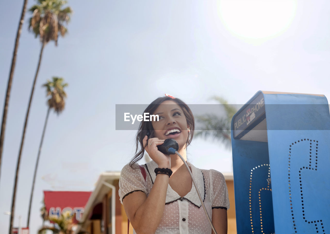 Young woman talking on pay phone while standing against sky