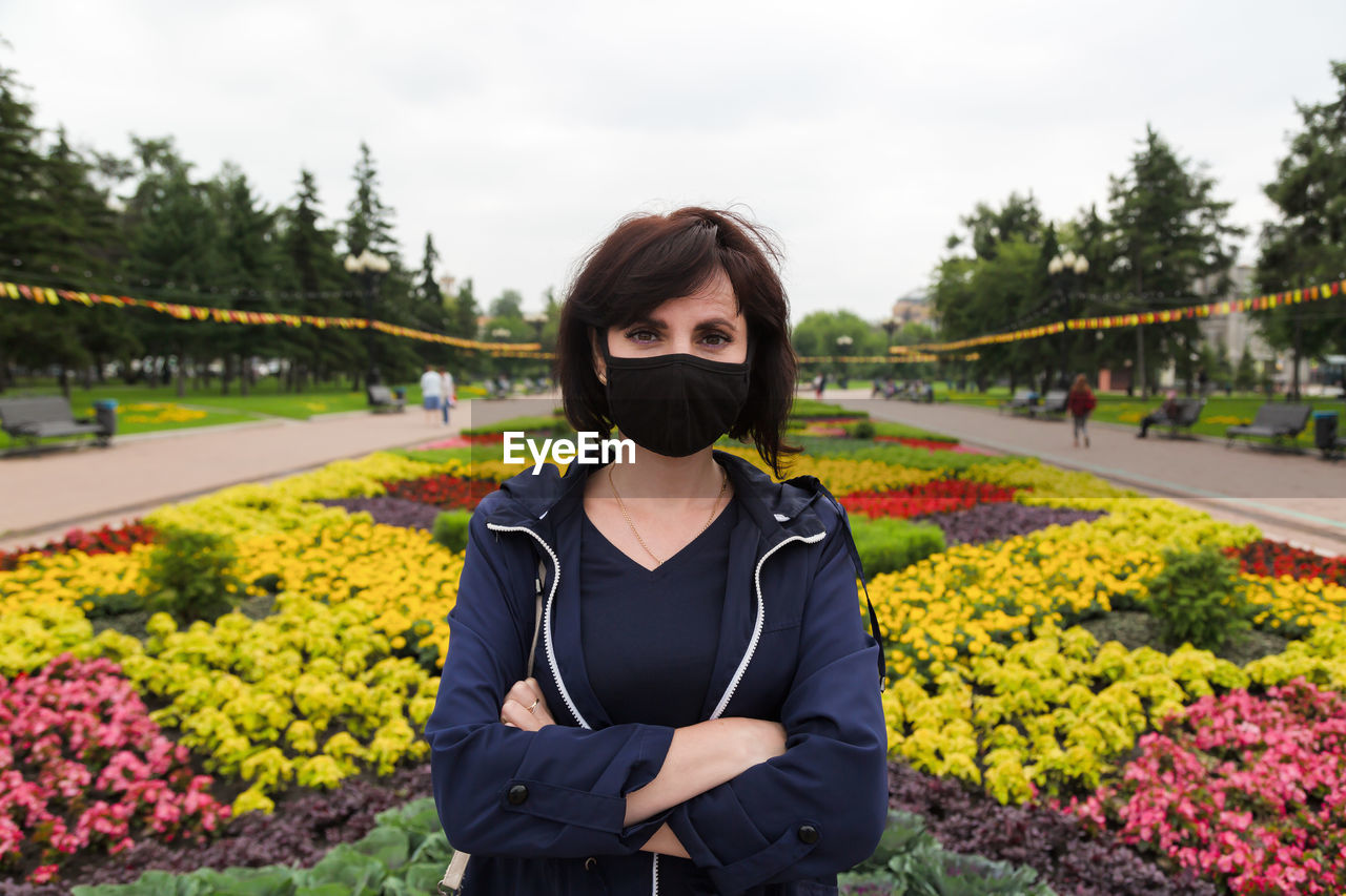 Portrait of a woman in a black protective mask on the background of a bright flower bed in the park.