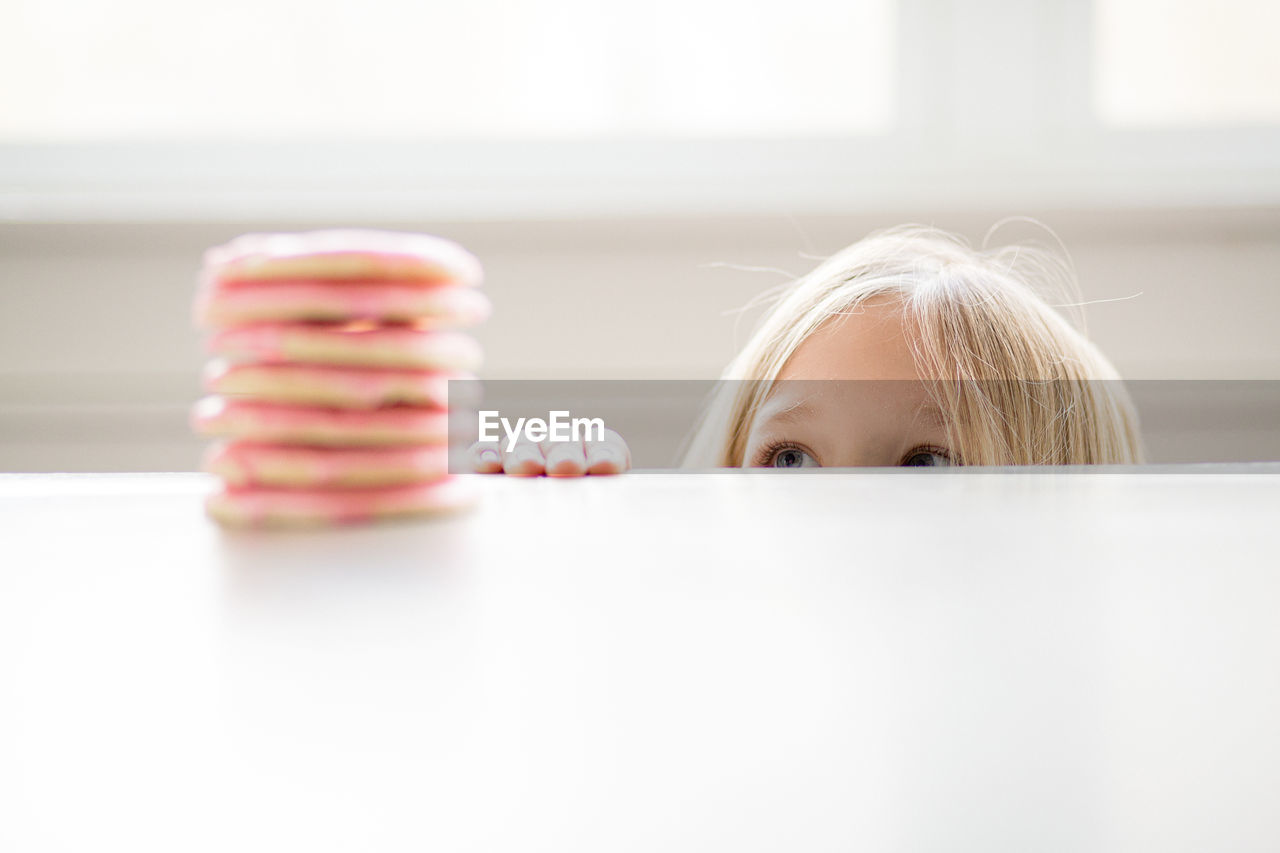 Blond girl peeking at stack of pink frosted sugar cookies on table