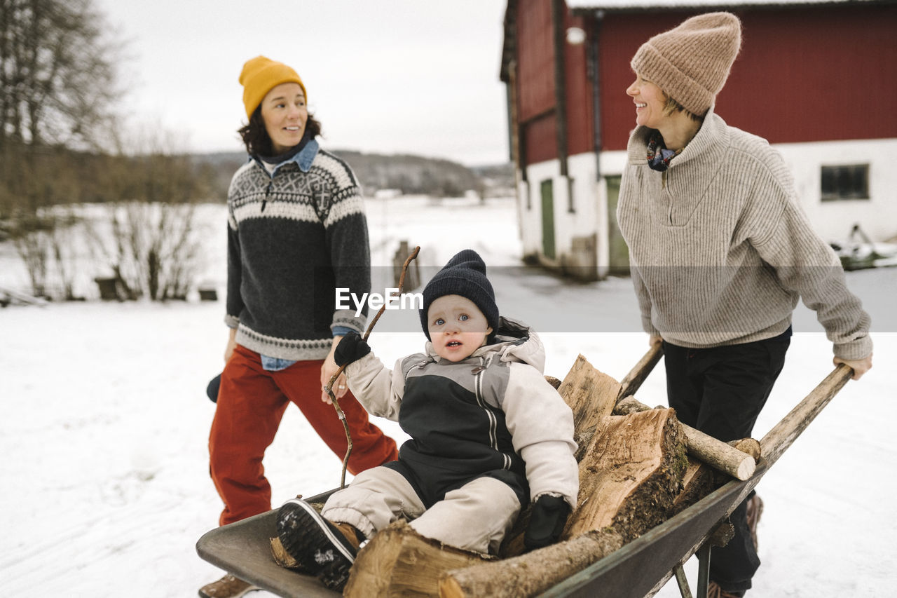 Smiling woman looking at girlfriend while carrying daughter in wheelbarrow during winter