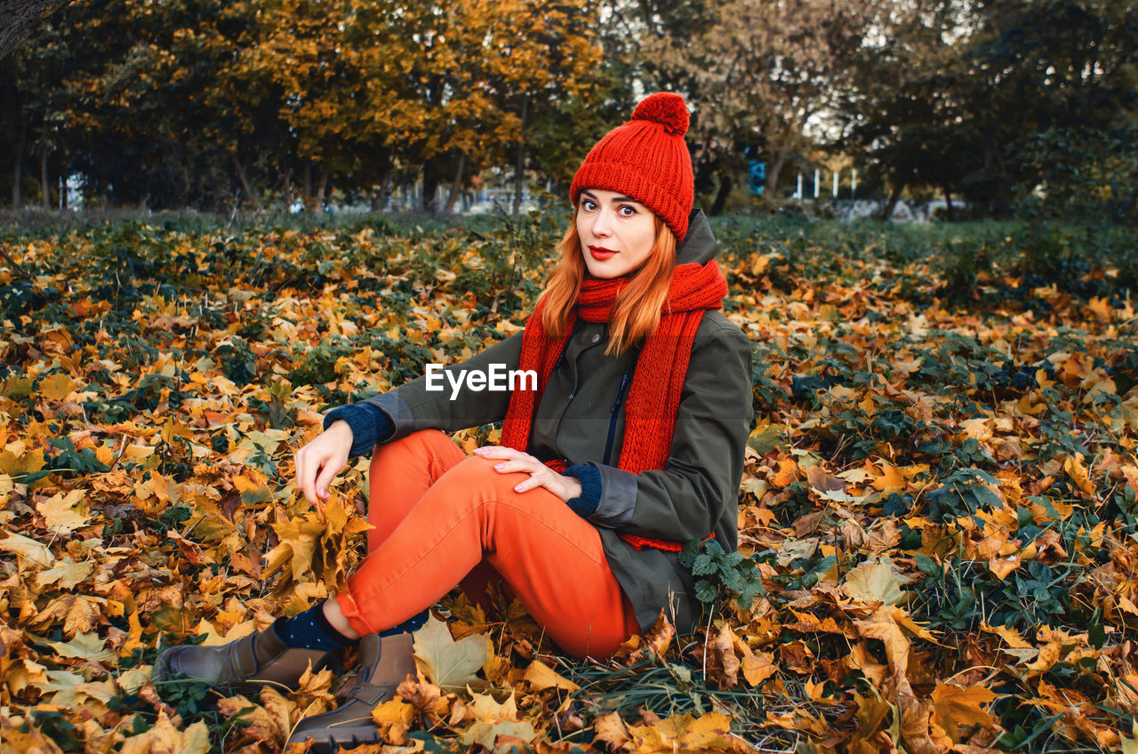 Autumn portrait of cute girl in orange warm knitted hat and orange scarf sitting in leafs 