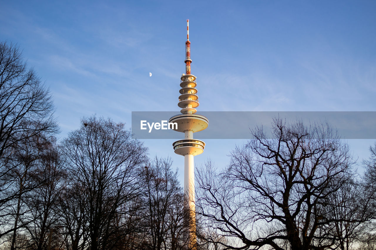 Low angle view of bare trees and communications tower against cloudy sky