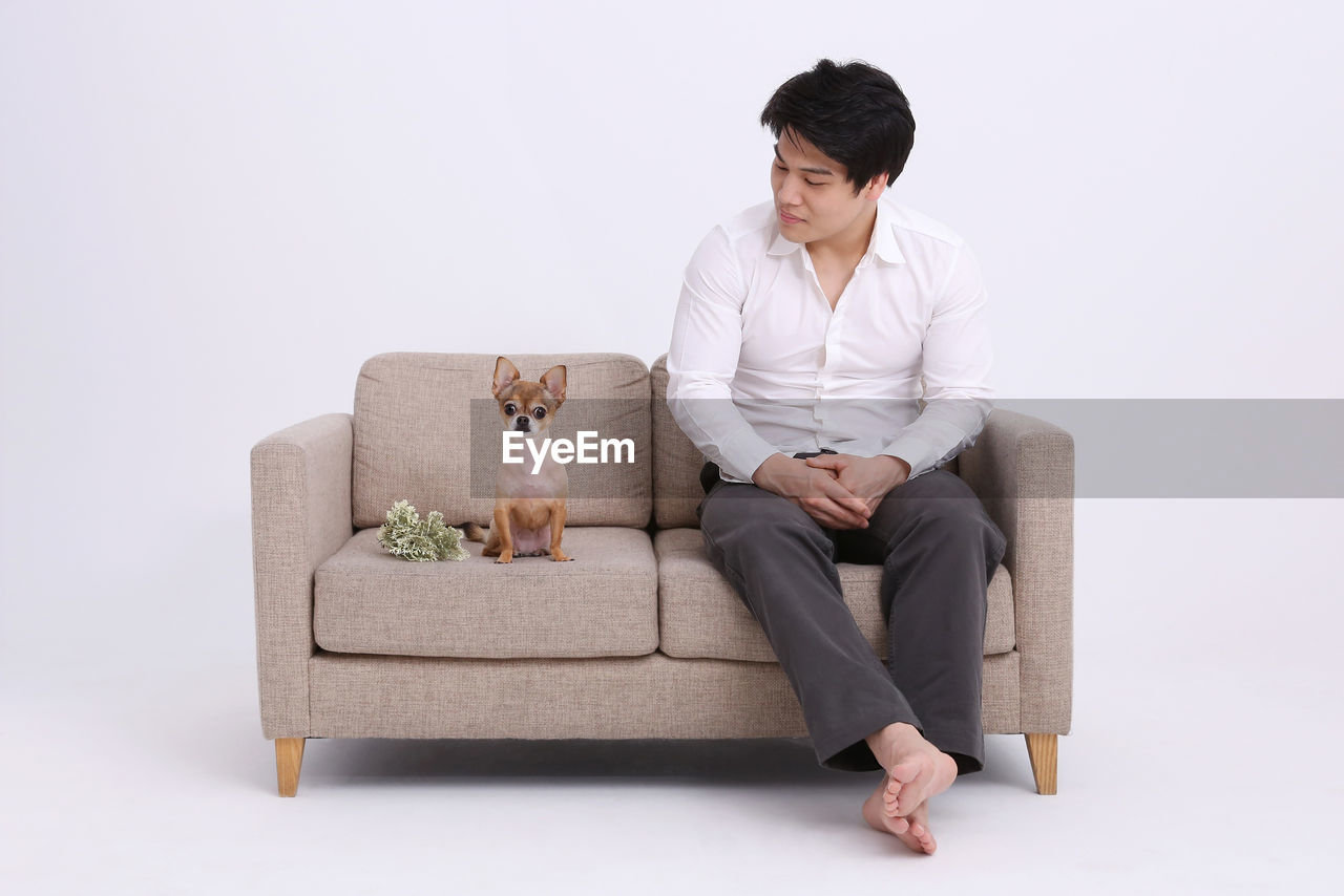 Full length of man sitting with dog on sofa against white background