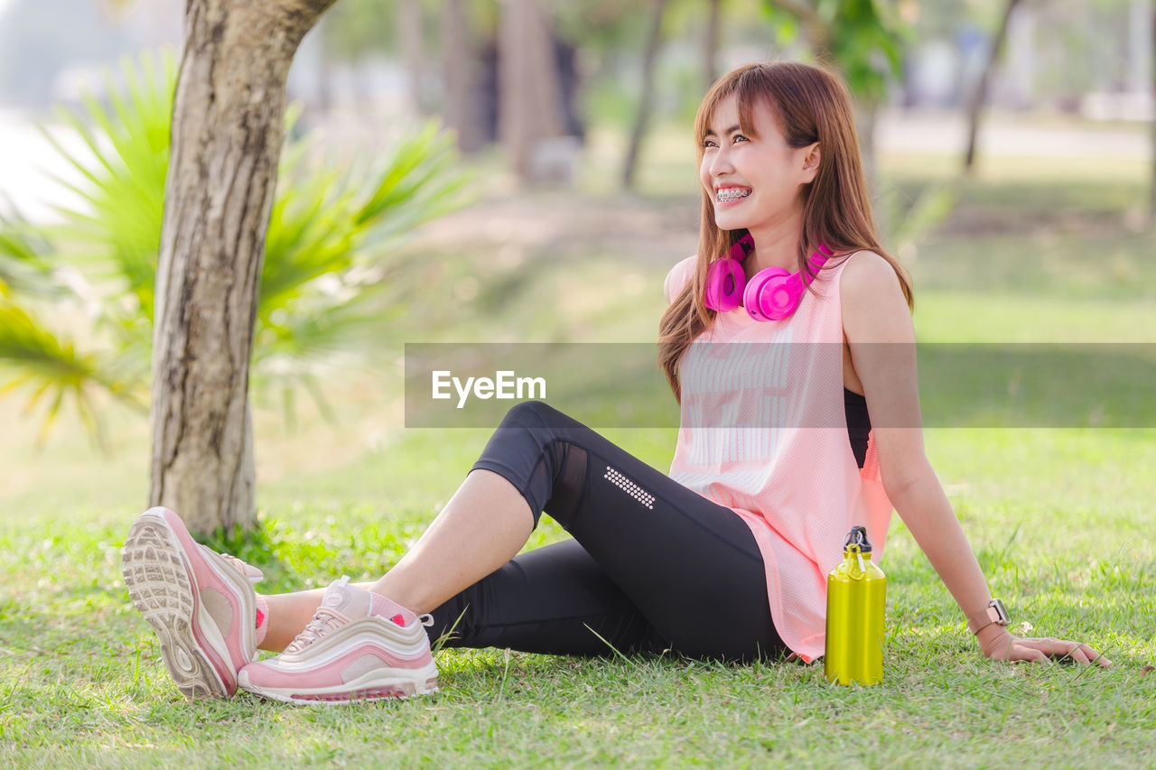 Side view of woman looking away while sitting on grass by water bottle