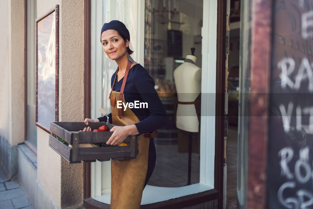 Female owner looking away while carrying vegetable crate at entrance of grocery store