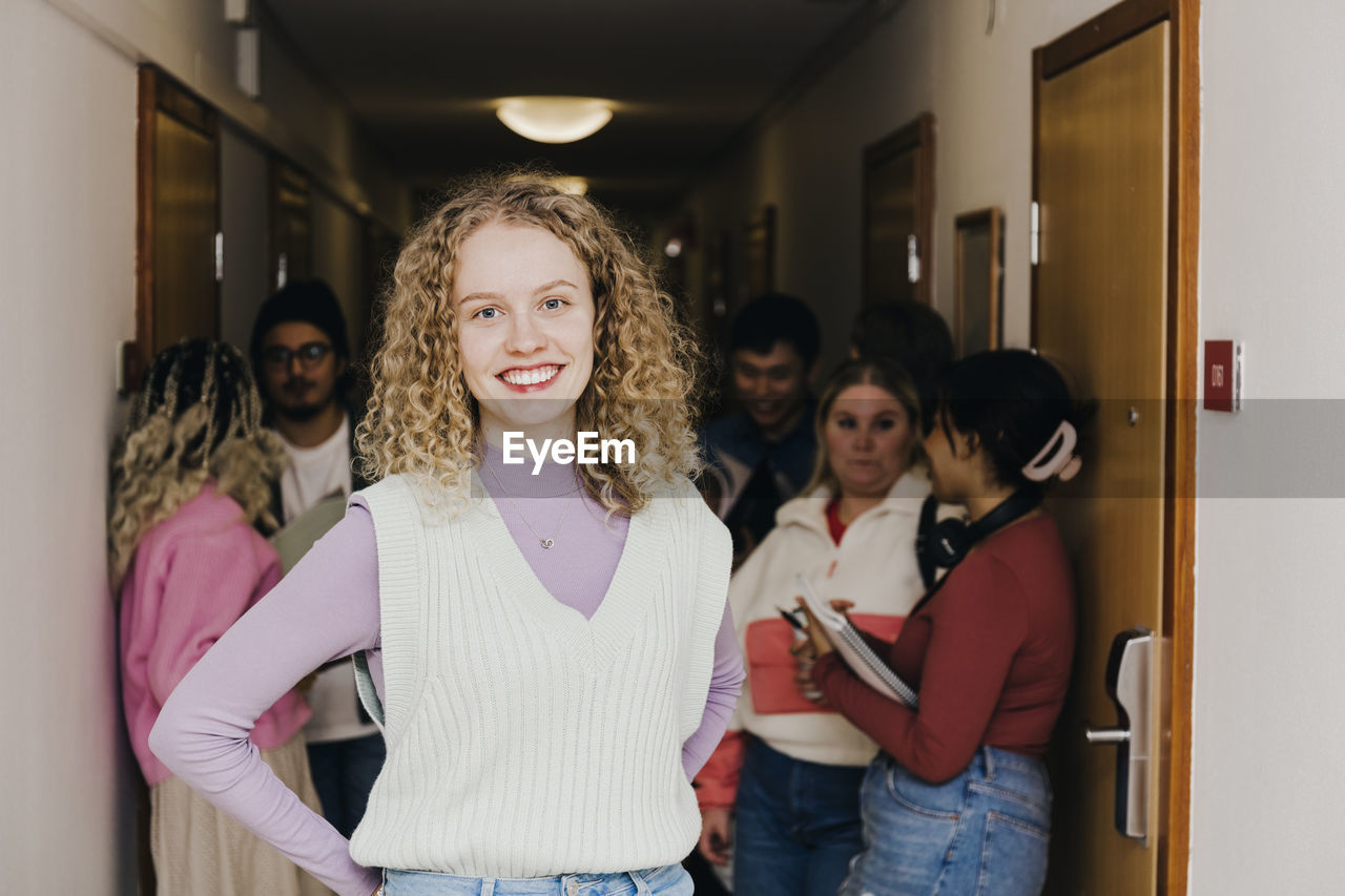 Portrait of smiling young woman with multiracial friends in background at corridor in college dorm