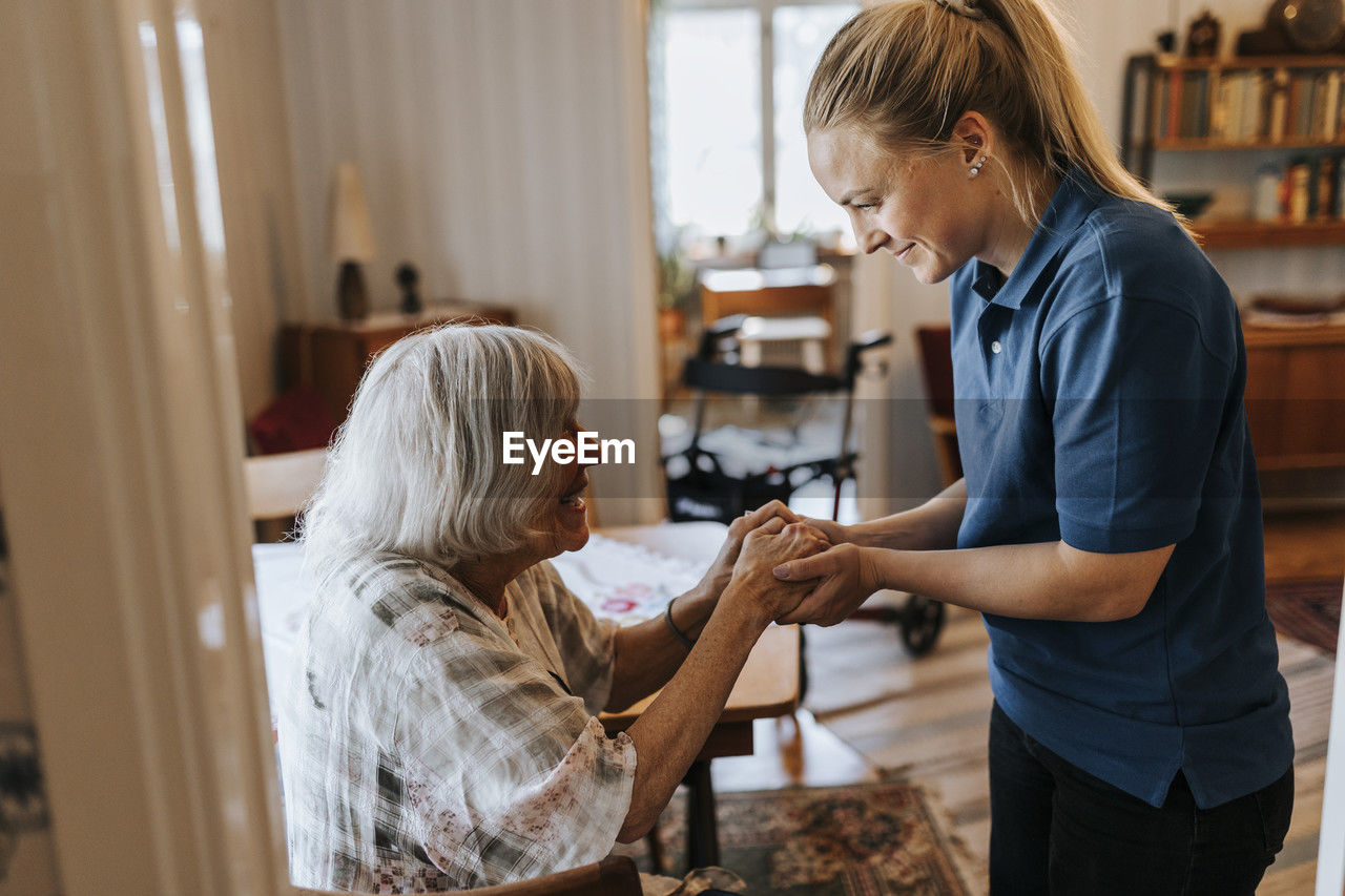 Smiling female healthcare worker holding hands of senior woman at home