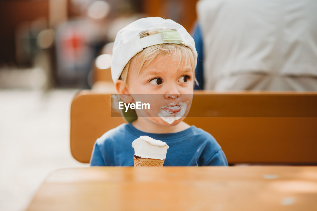 Young blonde boy with dirty face eating ice cream with a cap on