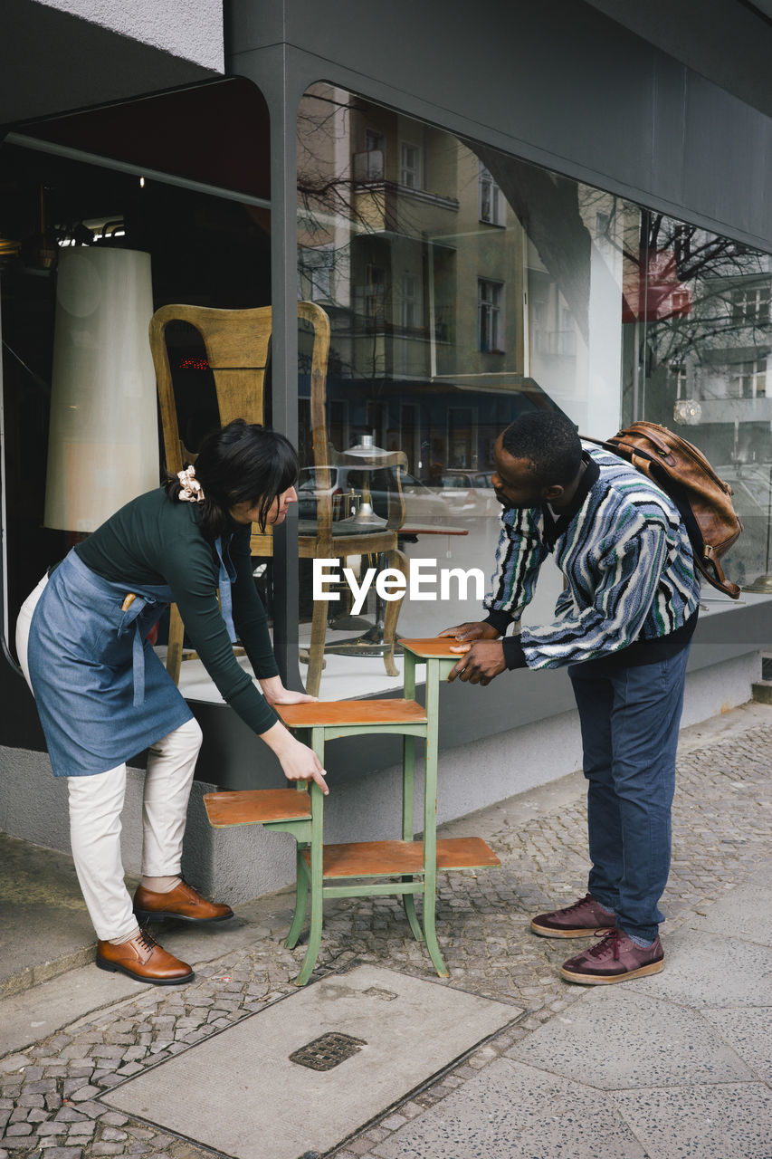 Female upcycling store owner showing furniture to male customer at sidewalk