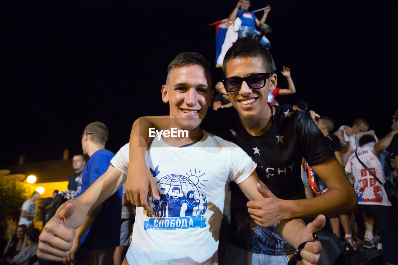 PORTRAIT OF HAPPY FRIENDS STANDING AT NIGHT DURING FESTIVAL