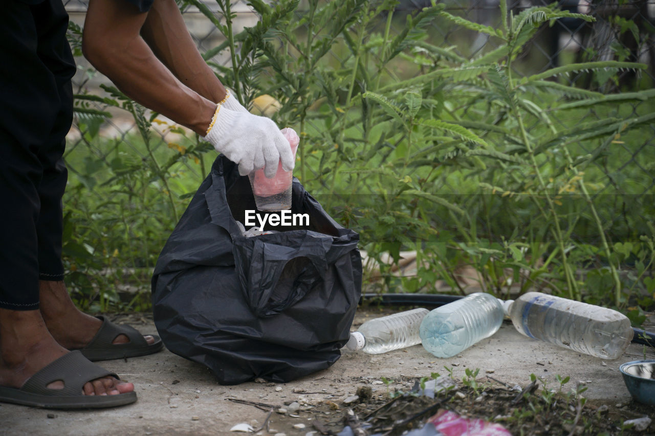 Low section of man holding glass over garbage bag by plants