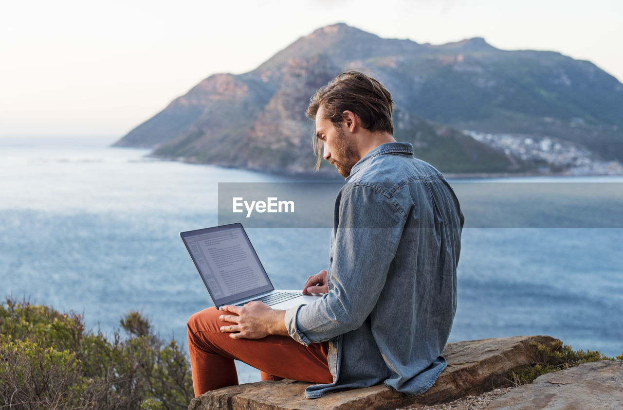 Side view of man using laptop on hill against mountain and sea