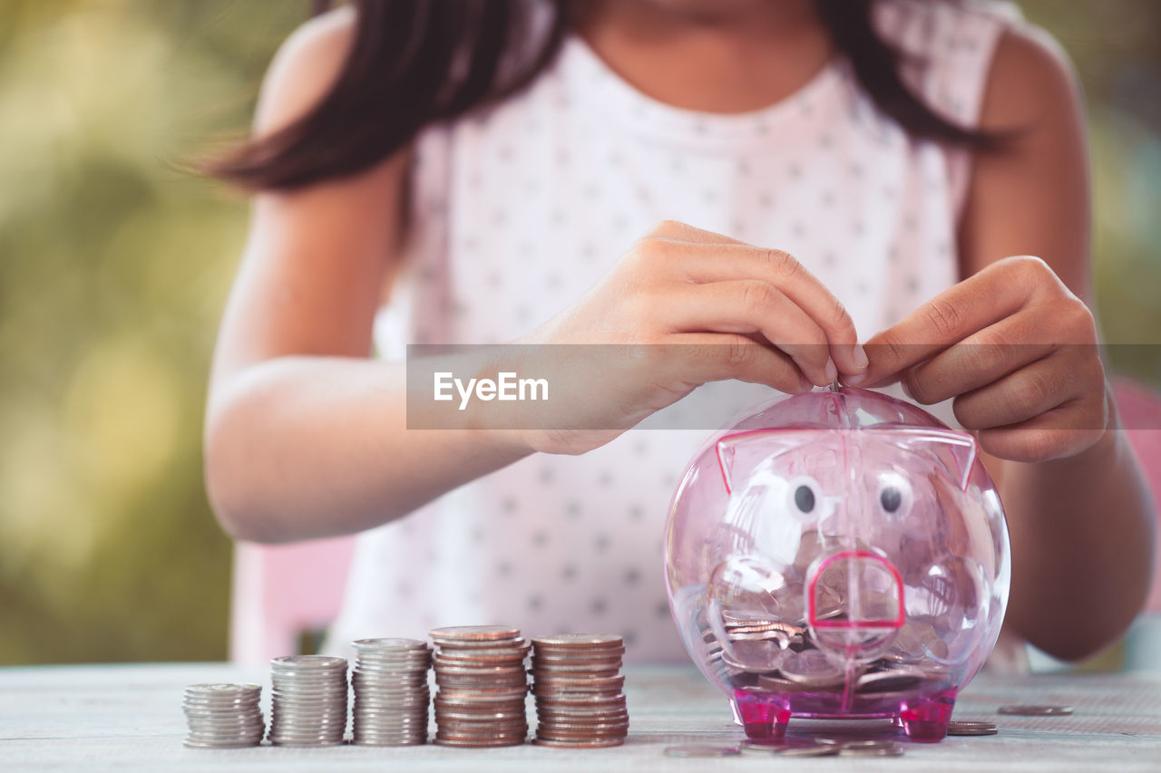 Midsection of girl putting coins in piggy bank on table