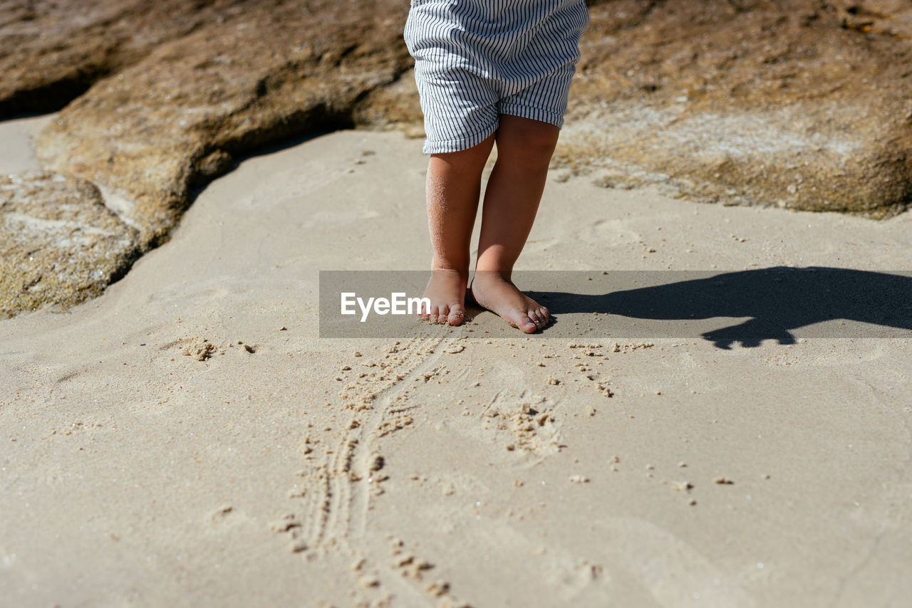 Faceless little child with bare feet standing on wet soft sand of shoreline with rocks in bright sunlight