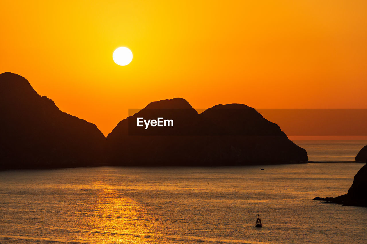 Scenic view of sea and silhouette mountains against orange sky