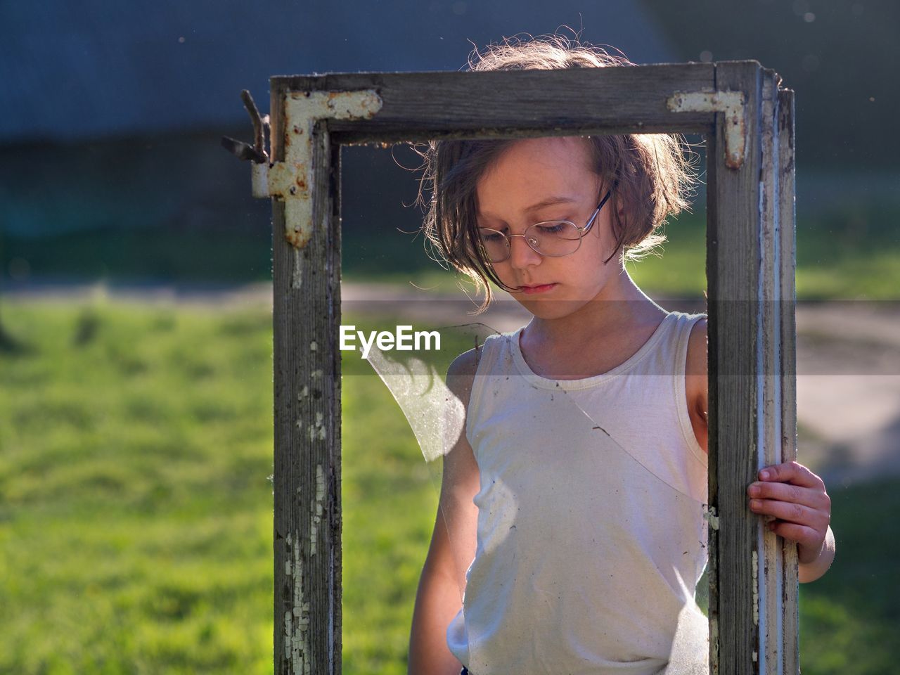 Sad boy holding broken old window while standing on grass