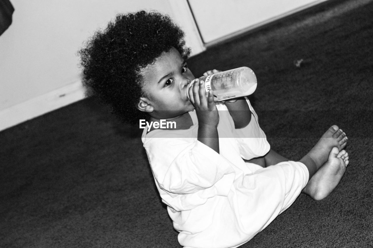 Tilt shot of baby boy drinking from milk bottle while sitting on carpet at home