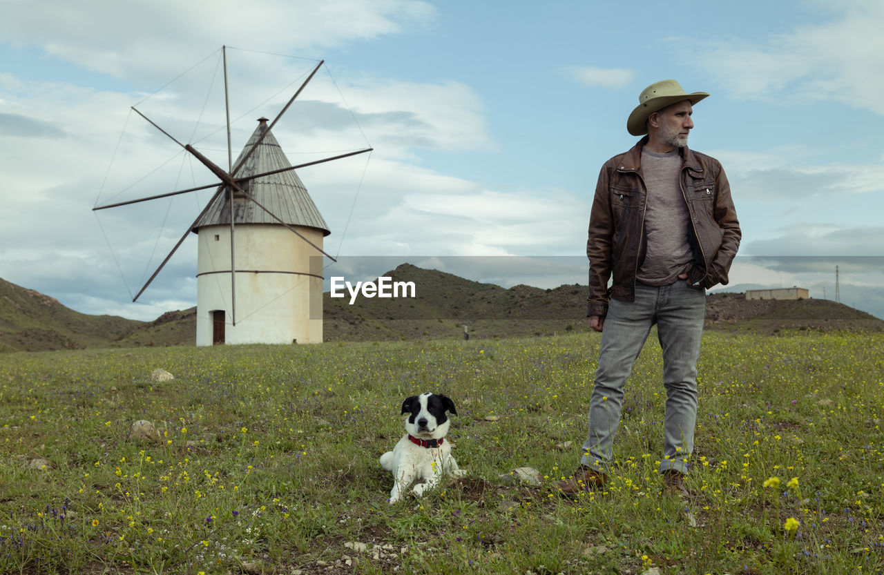 Adult man in cowboy hat and his dog standing on field with traditional windmill in background