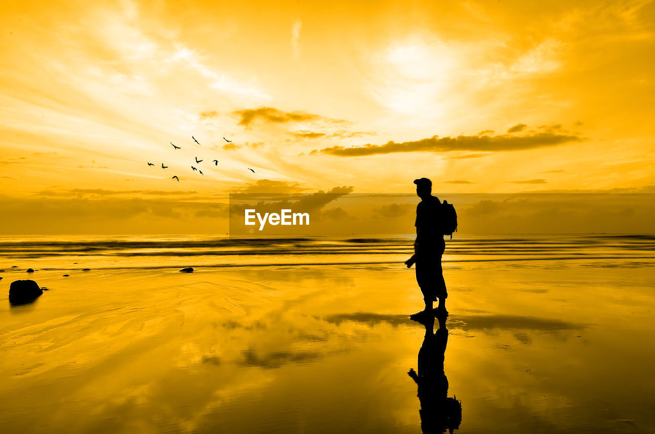 SILHOUETTE MAN STANDING ON SHORE AGAINST ORANGE SKY