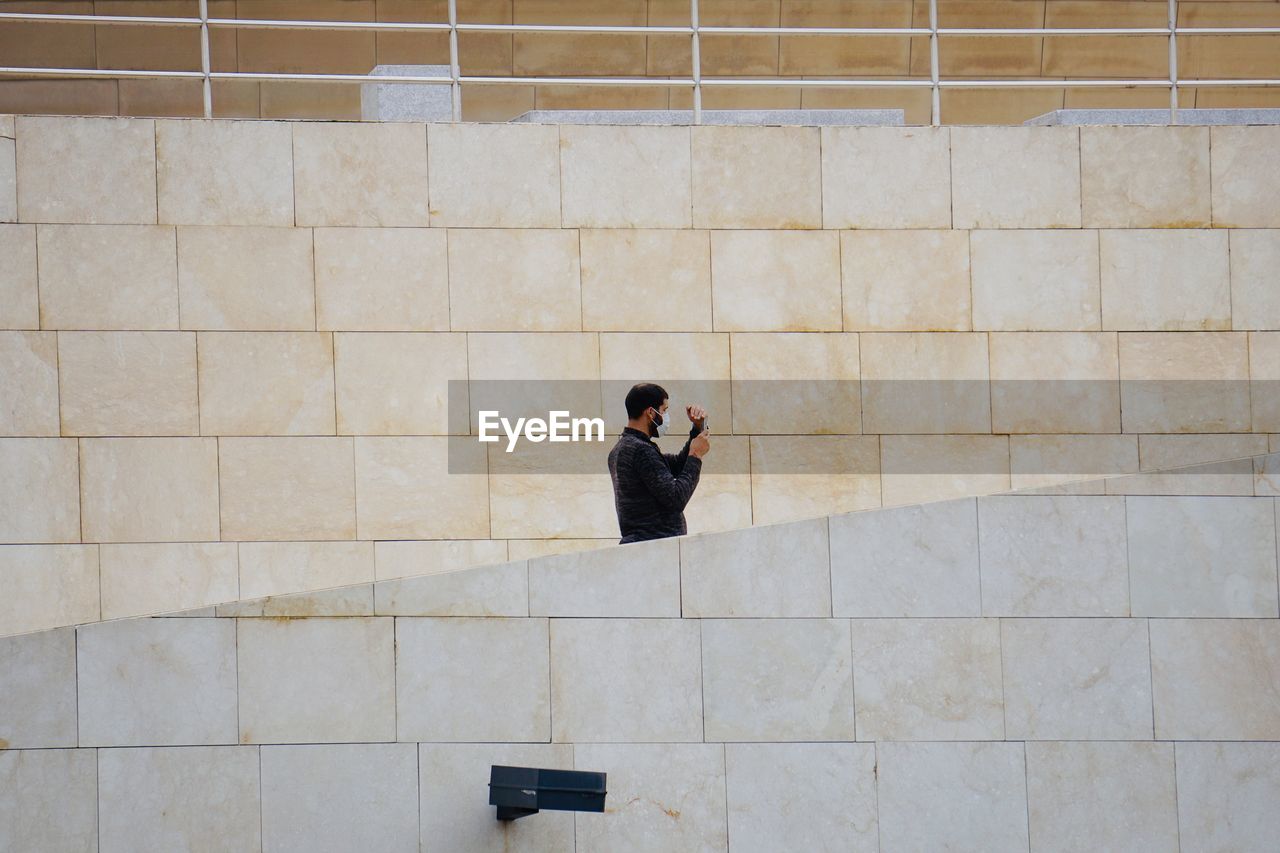 REAR VIEW OF MAN USING PHONE AGAINST WALL