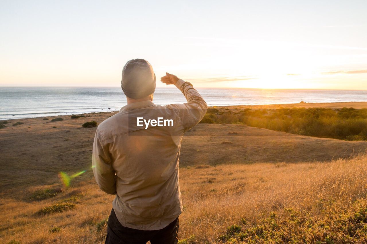 Rear view of man shielding eyes at shore against sky during sunset
