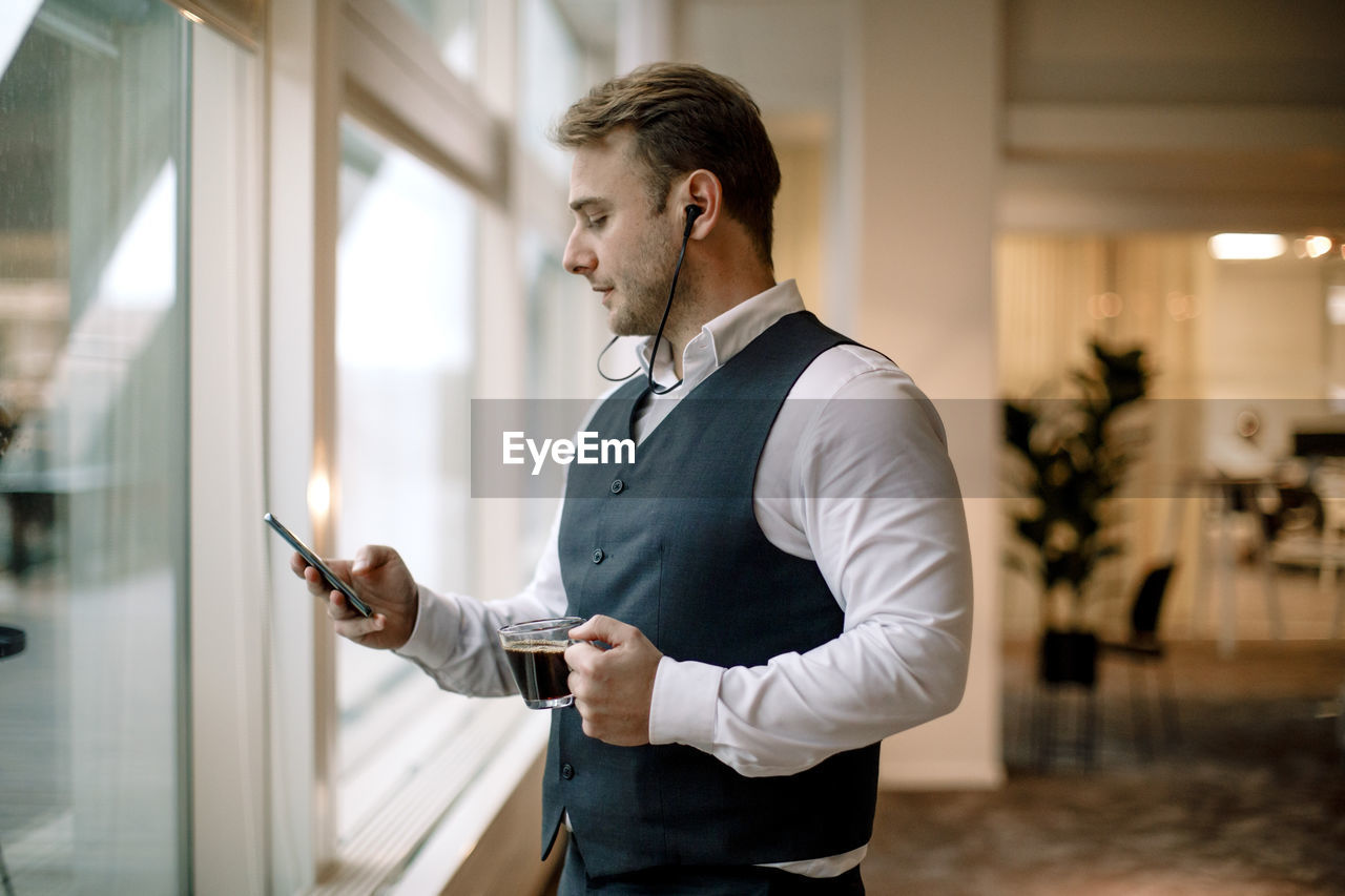 Businessman wearing headphones using mobile phone while having coffee at office