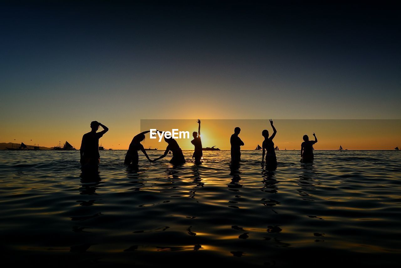 Silhouette friends standing in sea against clear sky during sunset