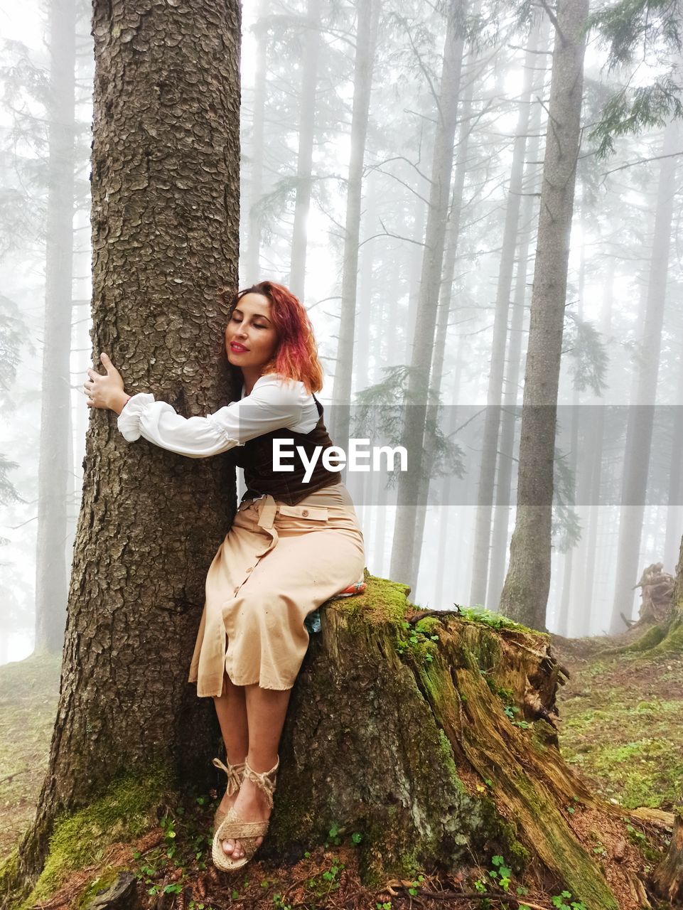 Full length portrait of woman standing by tree trunk in forest