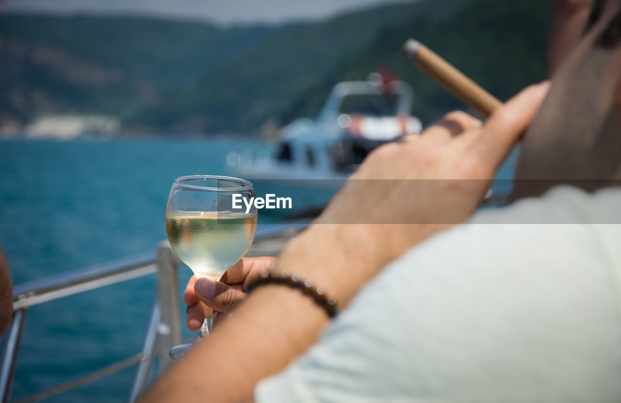 Close-up of hand holding wineglass over river against sky