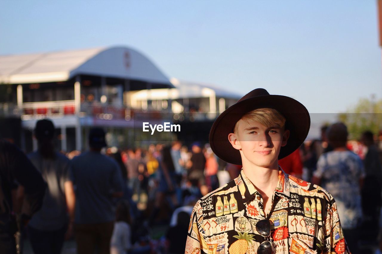 Portrait of young man wearing hat while standing in city against clear sky